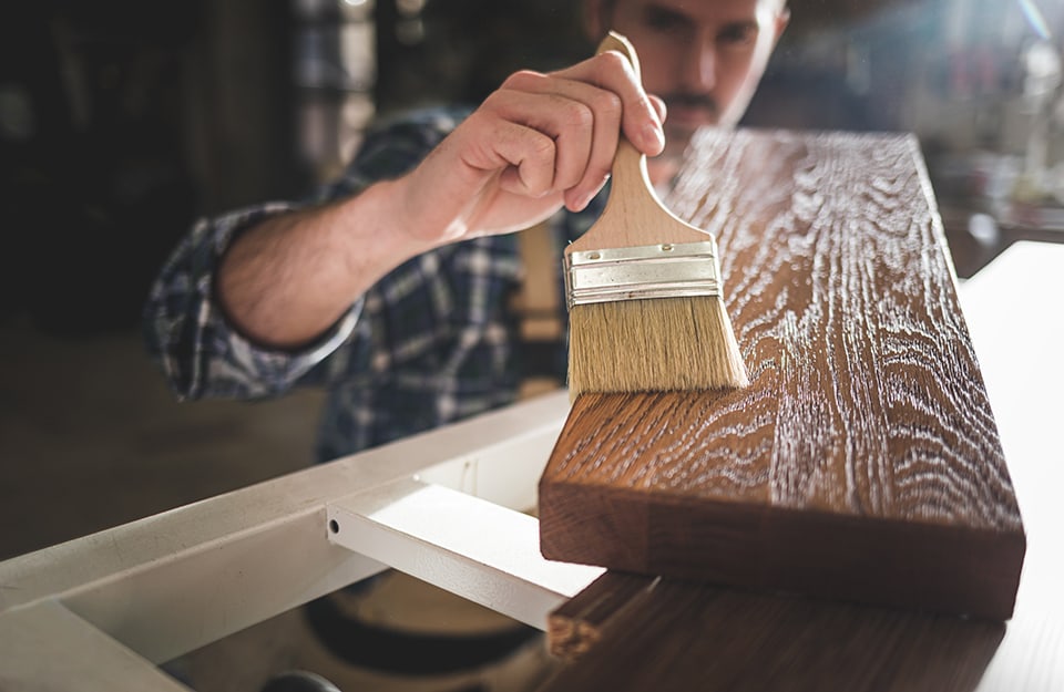 Un uomo con camicia a scacchi osserva un'asse di legno che sta trattando con del mordente per legno, steso con un pennello