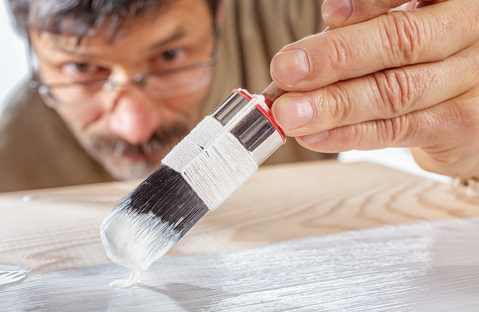 Un uomo con baffi e occhiali osserva da vicino mentre sta passando il fondo bianco per legno su un manufatto con un pennello giapponese