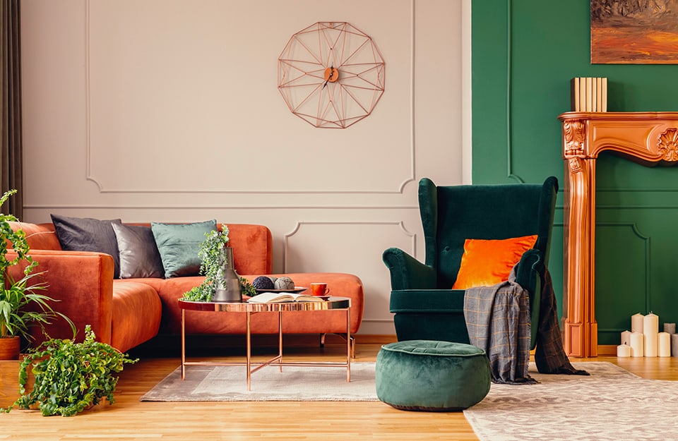 Un salotto arredato in stile eclettico, con sofà rosa-arancio e cuscini lucidi multicolore, una parete dipinta di verde e l'altra di colore neutro, pavimento a parquet, tavolino da caffè circolare in metallo e vetro, poltrona vintage verde scuro a effetto vellutato, pouf, piante, tappeti e la cornice in legno di un camino con delle candele al posto del fuoco