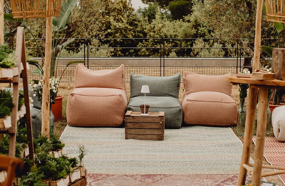 Vista su un giardino in stile boho-chic con pouf in colori tenui e tessuti lucidi, grandi tappeti stesi a terra, strutture in legno, grandi piante, un tavolino da caffè realizzato con una cassetta della frutta